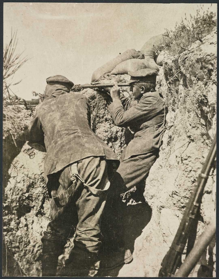 Two soldiers in a trench, one is looking through a telescope while the other the aims a rifle.