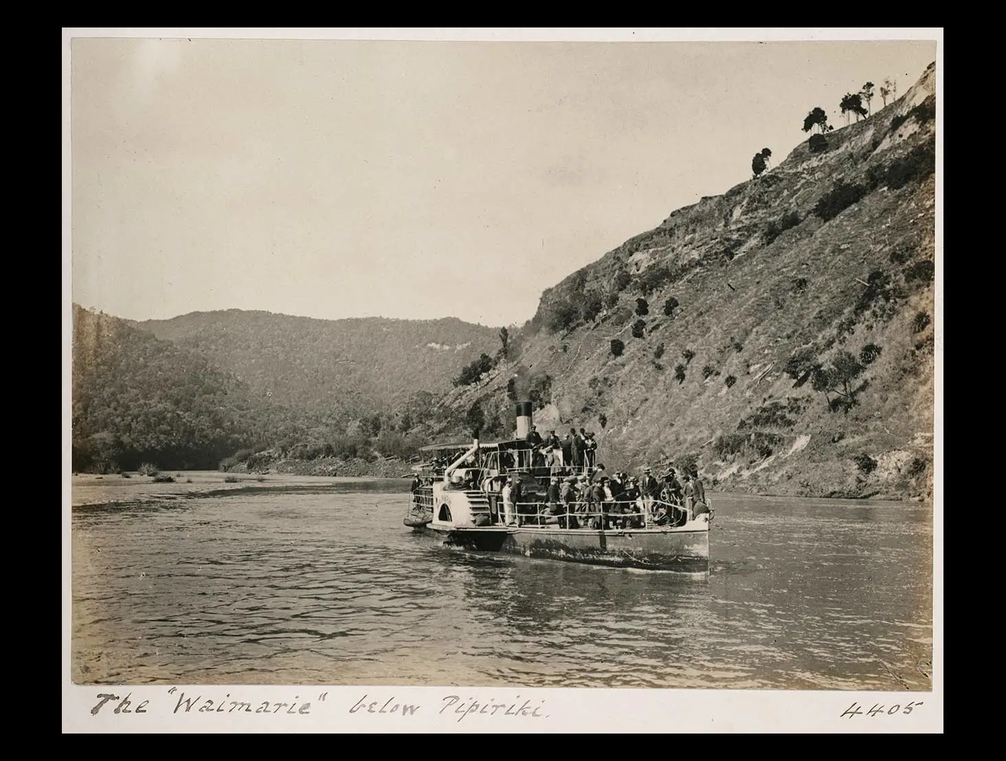 Black and white photo of the paddle steamer ‘Waimarie’ sailing on the Whanganui River with steep hills and bush in the background.