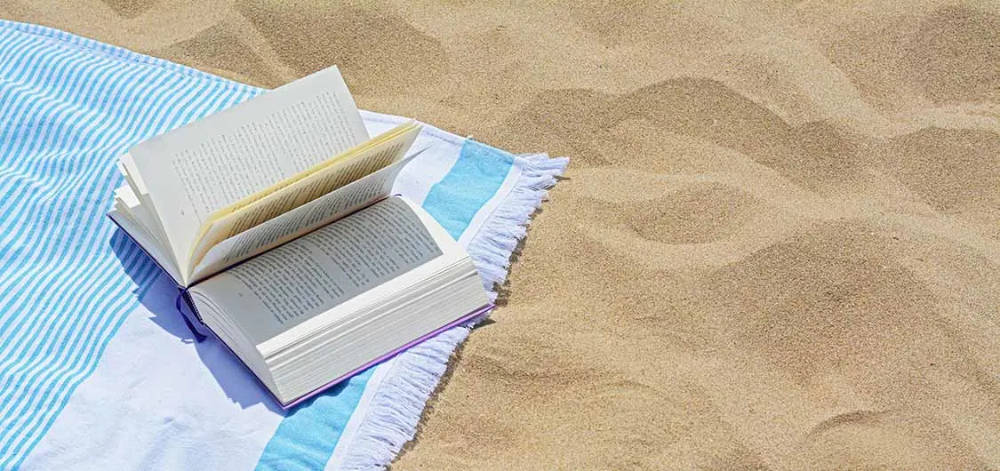 An open book resting on a beach towel placed on sand.