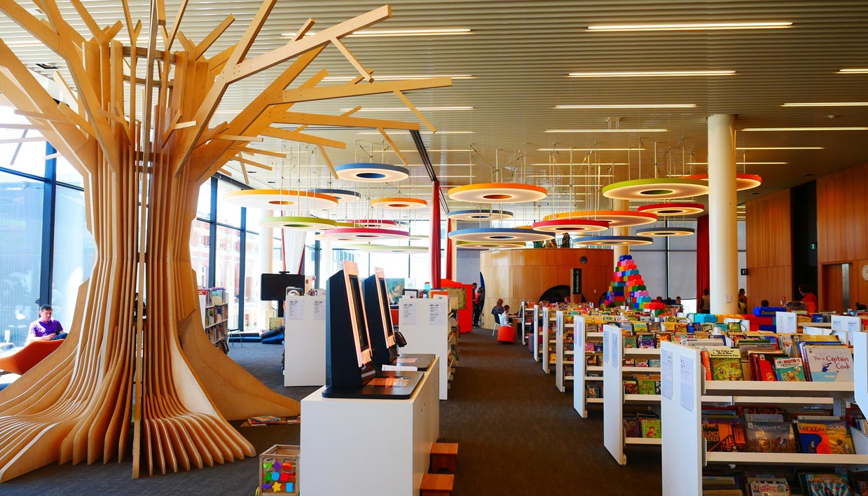 Photo of the children's area at Tūranga (Christchurch Central Library) with wooden tree-like sculpture, rows of shelving, toys, search screens, bright coloured ring-shaped ornaments hanging from ceiling.