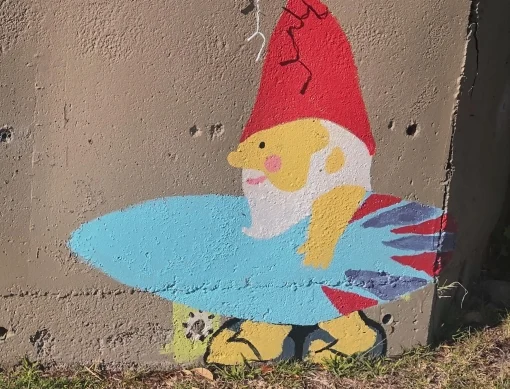 A gnome wearing a red hat and holding a surfboard has been painted on a concrete wall. 