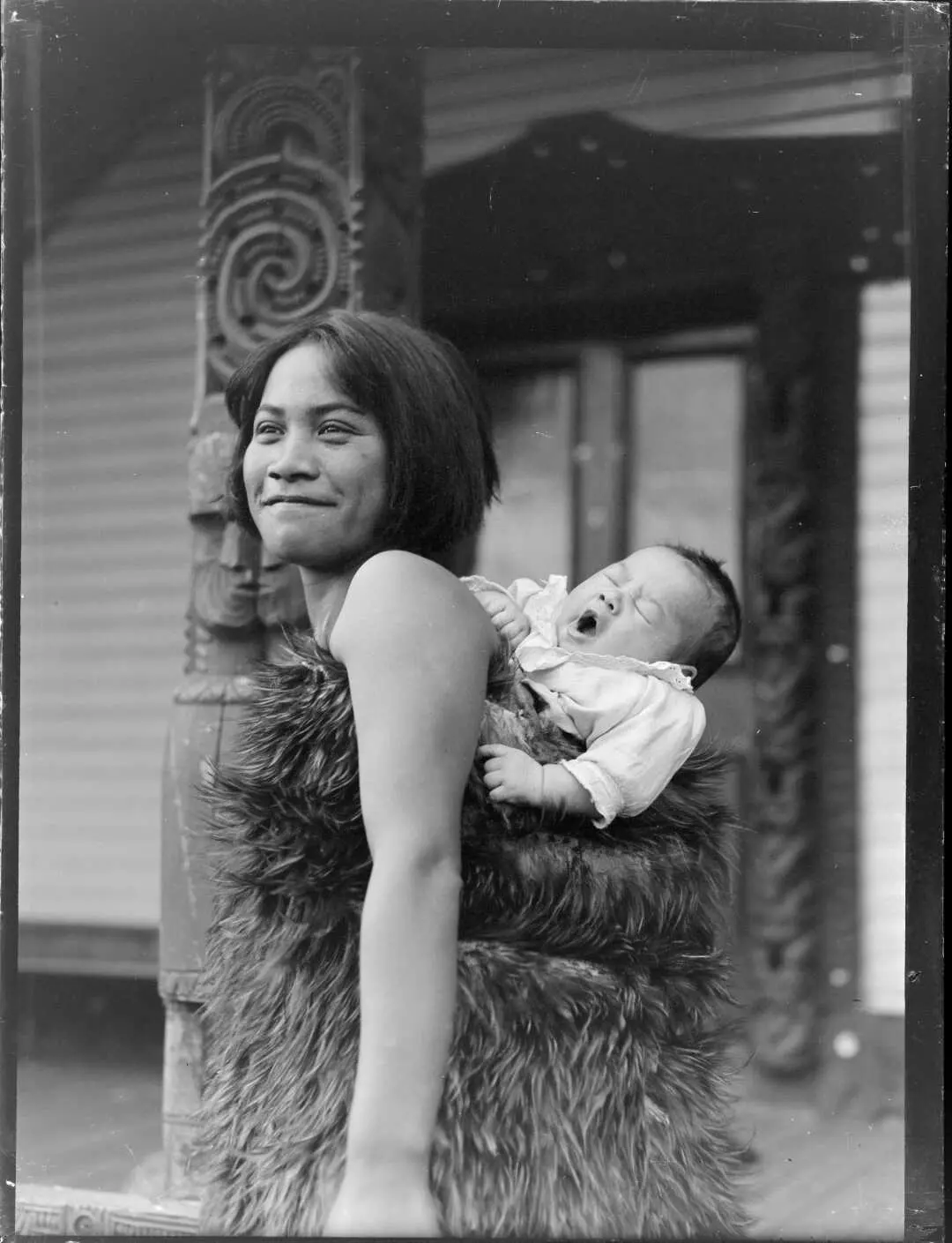 Maori woman carrying a young child on her back wrapped in a feather cloak. Ref: WA-12537-G