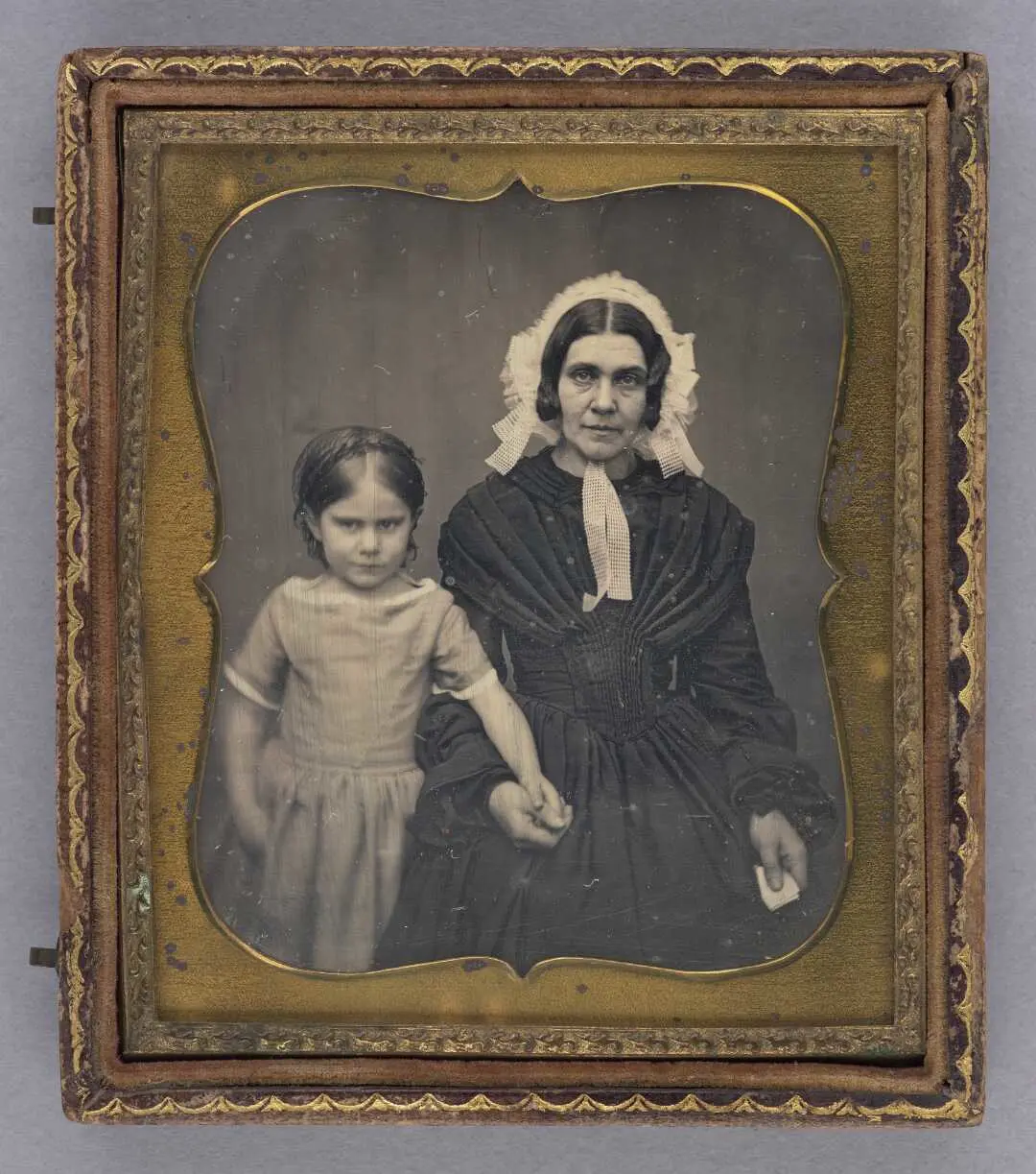 A portrait of a  tired looking woman and recalcitrant looking young child. They are holding hands.  Picture is in a small gold frame. 