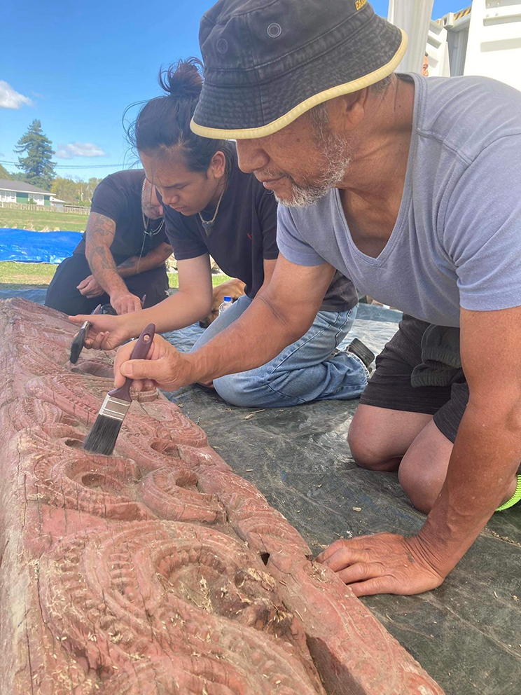 Two Māori men kneeling on the ground work together carefully brushing carvings that have been covered in silt. 
