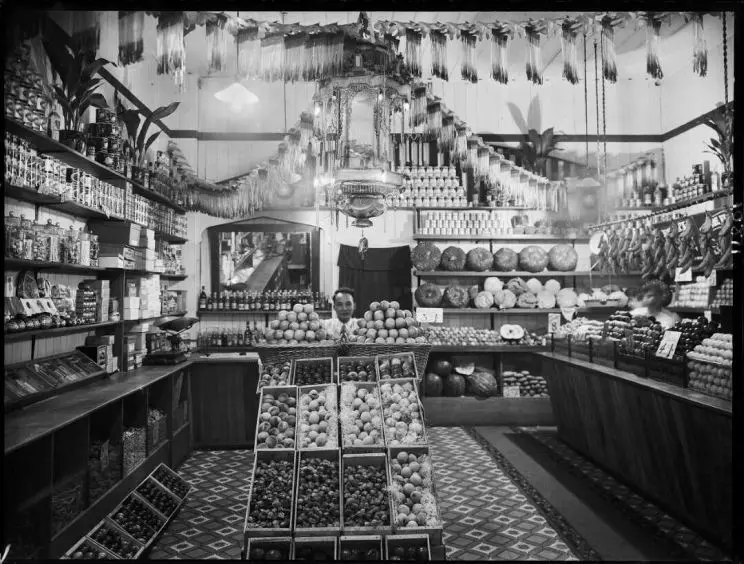 Greengrocery shop interior, probably in the Taranaki region. Shows a Chinese shopkeeper in the centre of the store, standing behind baskets of apples and boxes containing peaches, cape gooseberries, and other fruits. More produce lines the shelves on the walls. A Chinese lantern, and paper streamers, hang from the ceiling.