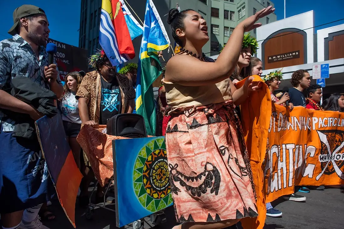 Colour photo of Pacific Climate Warriors supporting the Schools 4 Climate protest on a Wellington street. Supporters wear clothes with Pacific patterns and hold Pacific Islands flags and a large banner.