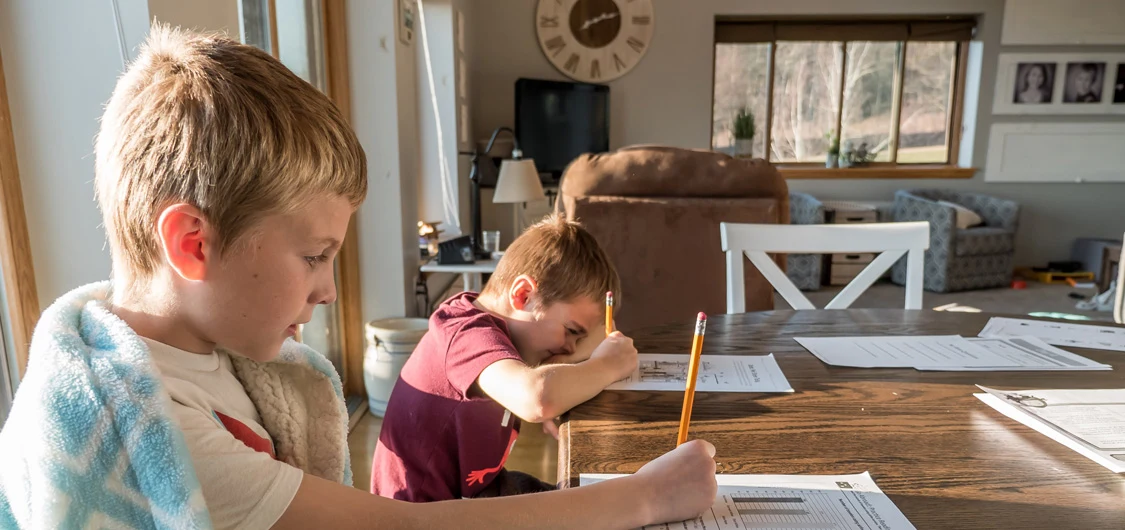 2 boys sitting at a dining table doing schoolwork. The boy in the foreground has a blanket wrapped around him.