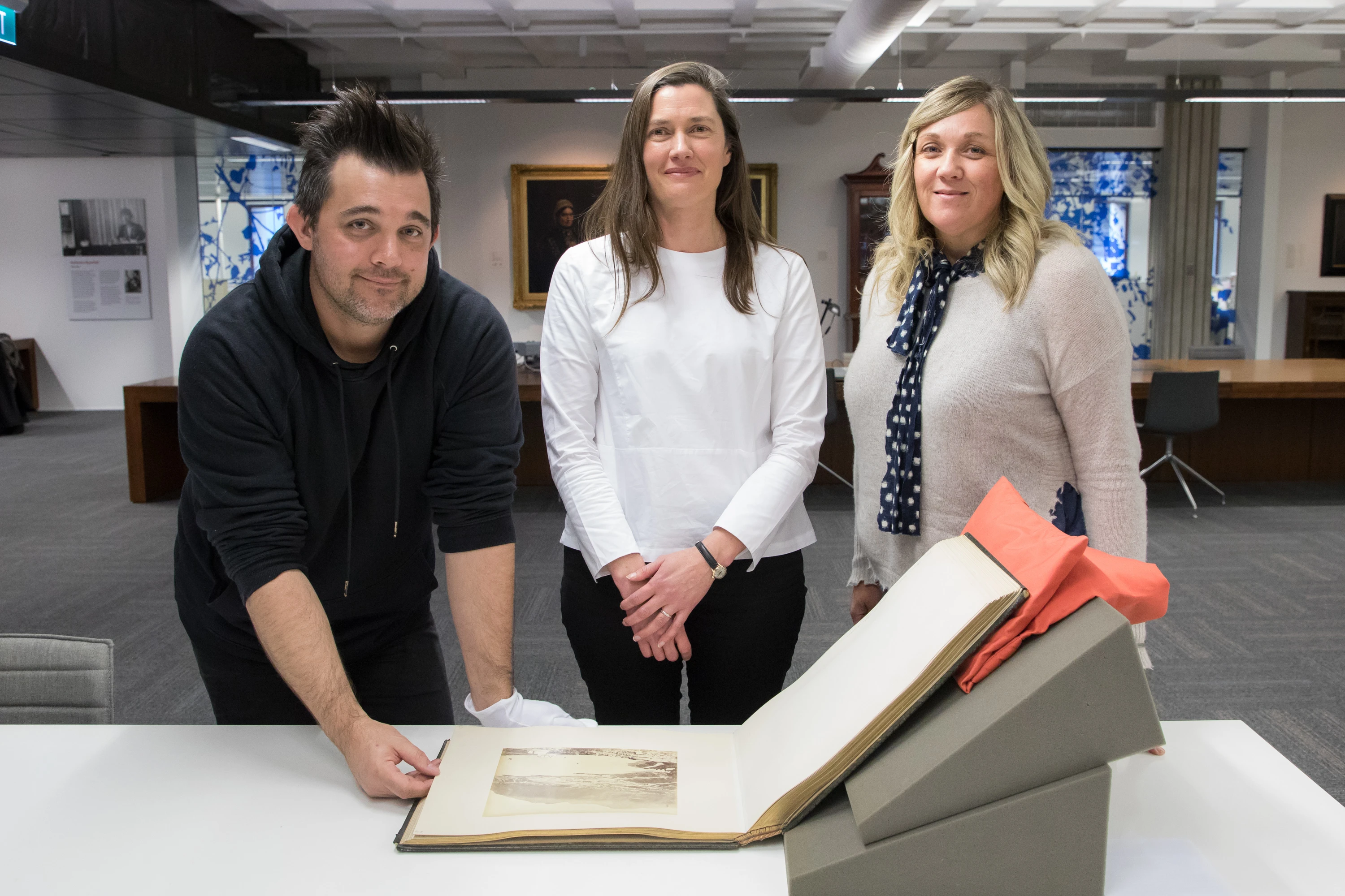 The authors of the blog stand with Photographic curator and on the table in front of them sits a large bound volume with text and images. 