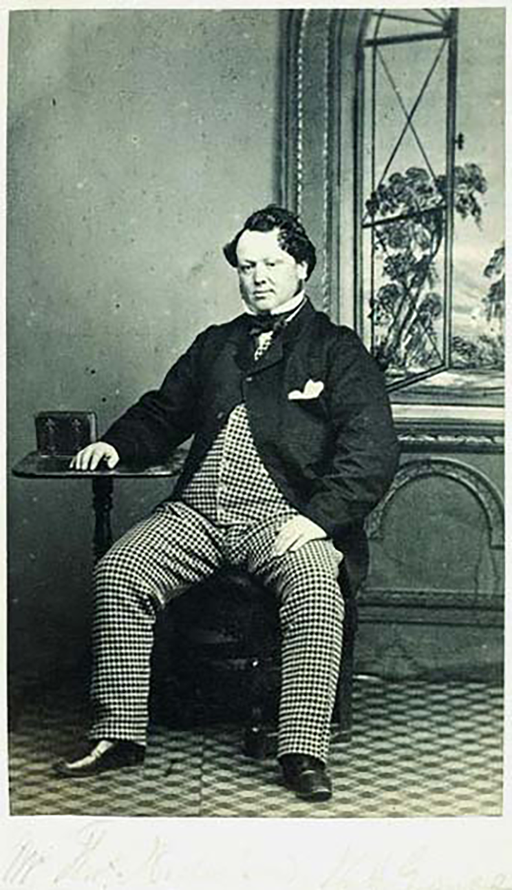 Victorian era photograph of a man in a checked suit sitting with his hand relaxing on a table. There is a picture of an open window behind him. 