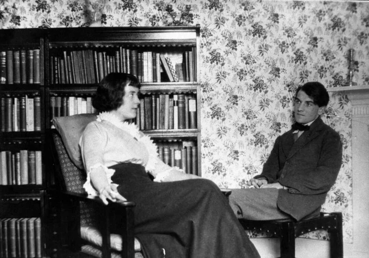 A black and white photo of a man and woman seated in a room as if in conversation with a bookshelf full of books in the background.