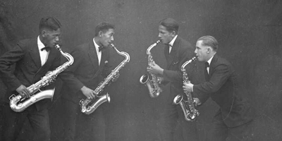 Four Māori men playing saxaphones. They are all wearing dark suits. 