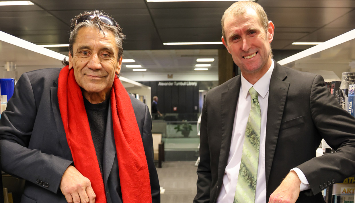 Inaugural Te Awhi Rito New Zealand Reading Ambassador Ben Brown with new ambassador Alan Dingley in a library.