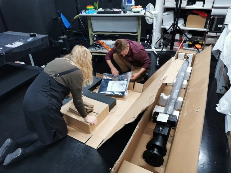 Two staff members opening the large cardboard box on the ground, revealing contents inside.