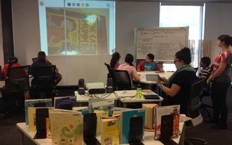 Children in a classroom staring at a book cover on a large screen, alongside various children's books on a desk.