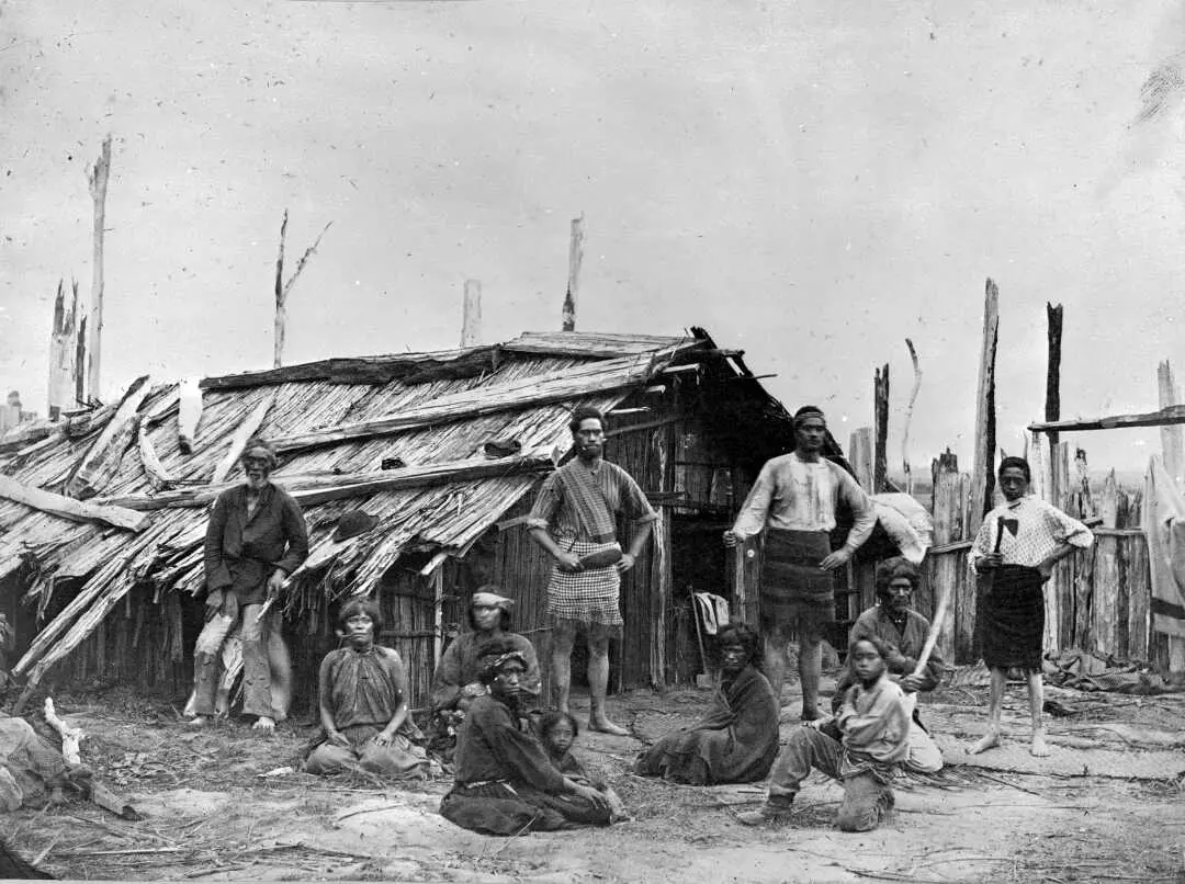 Shows both men and women standing and seated in front of a wooden whare.