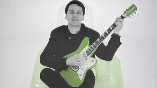 Shows a photograph of a smiling Martin Phillips holding a Charvel electric guitar and seated in a see-through blow-up armchair.
