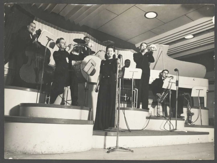 Black and white photo shows Lauri Paddi on stage with her band behind her, playing their instruments.