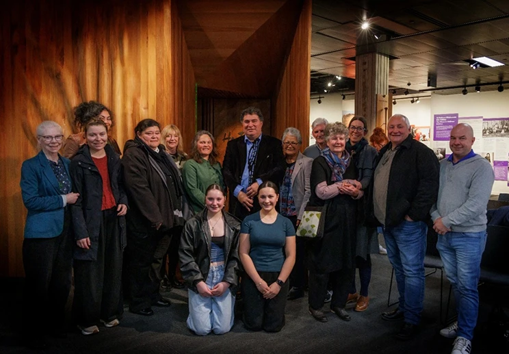 A diverse group of people posing together outside He Tohu, with wooden panels and exhibition displays visible in the background.