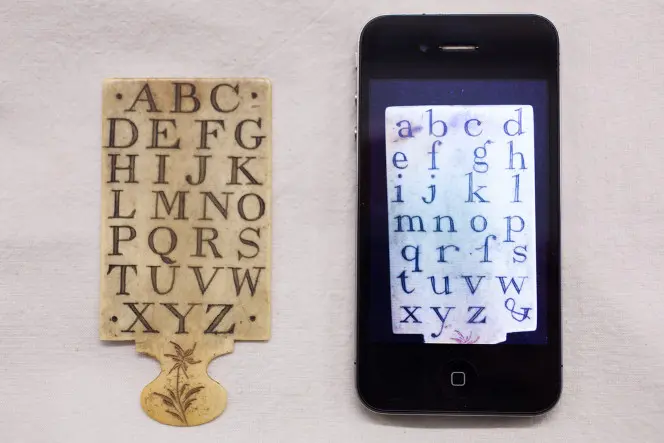 Hornbook showing uppercase alphabet, next to a cellphone. The cellphone is showing the reverse side with lowercase letters.