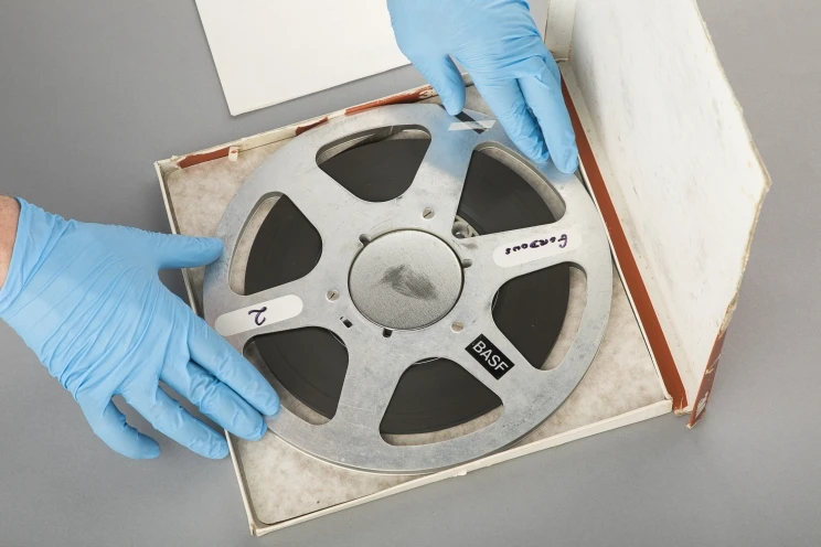 A reel of magnetic tape sitting inside its cardboard box with the technicians hands shown wearing blue rubber gloves.