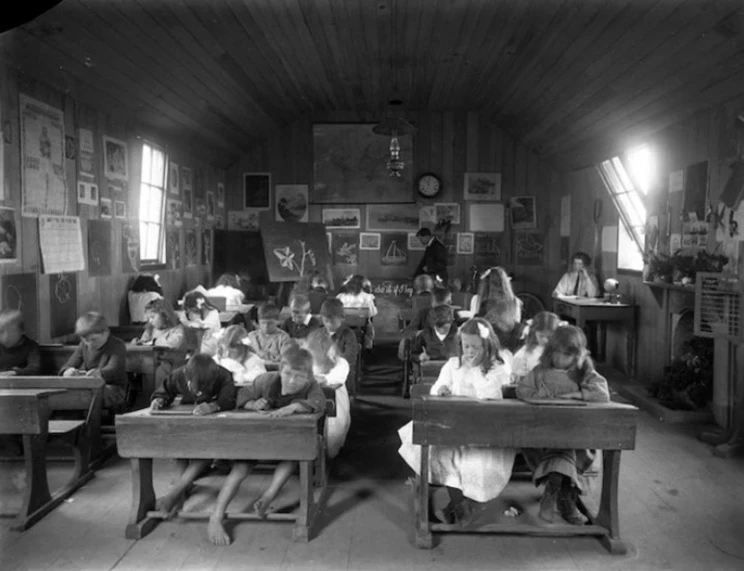 Black and white photo of children writing at desks in rows in an early 19th century classroom.
