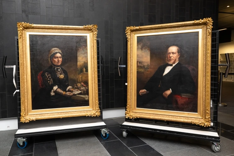 Two large, framed portraits of a man and a woman in formal dress, sit on wheeled supports.