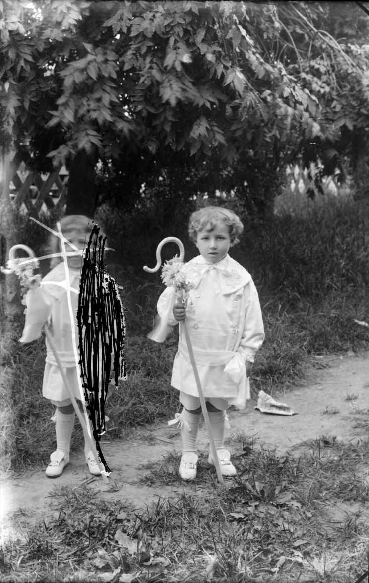 Two children stand side by side in identical white costumes, each holding a cane with flowers on the handle, once child has been partially scratched out of the negative.