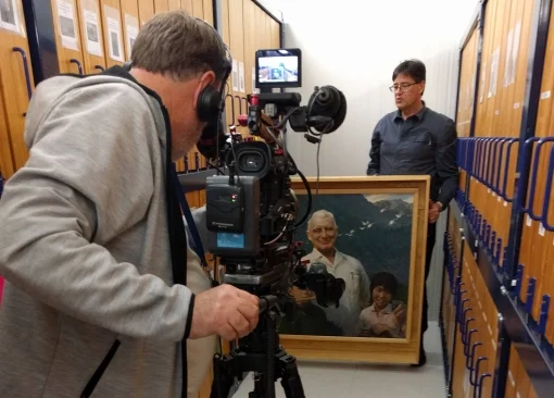 A  man points a video camera at a man standing behind a painting.