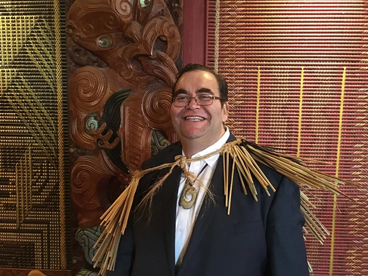 A photo of a Māori man standing in front of carved poupou and tukutuku panels.