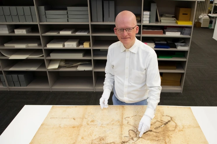 A man in a white button-down shirt and white gloves stands pointing at a certain location on an old, yellowing, map.