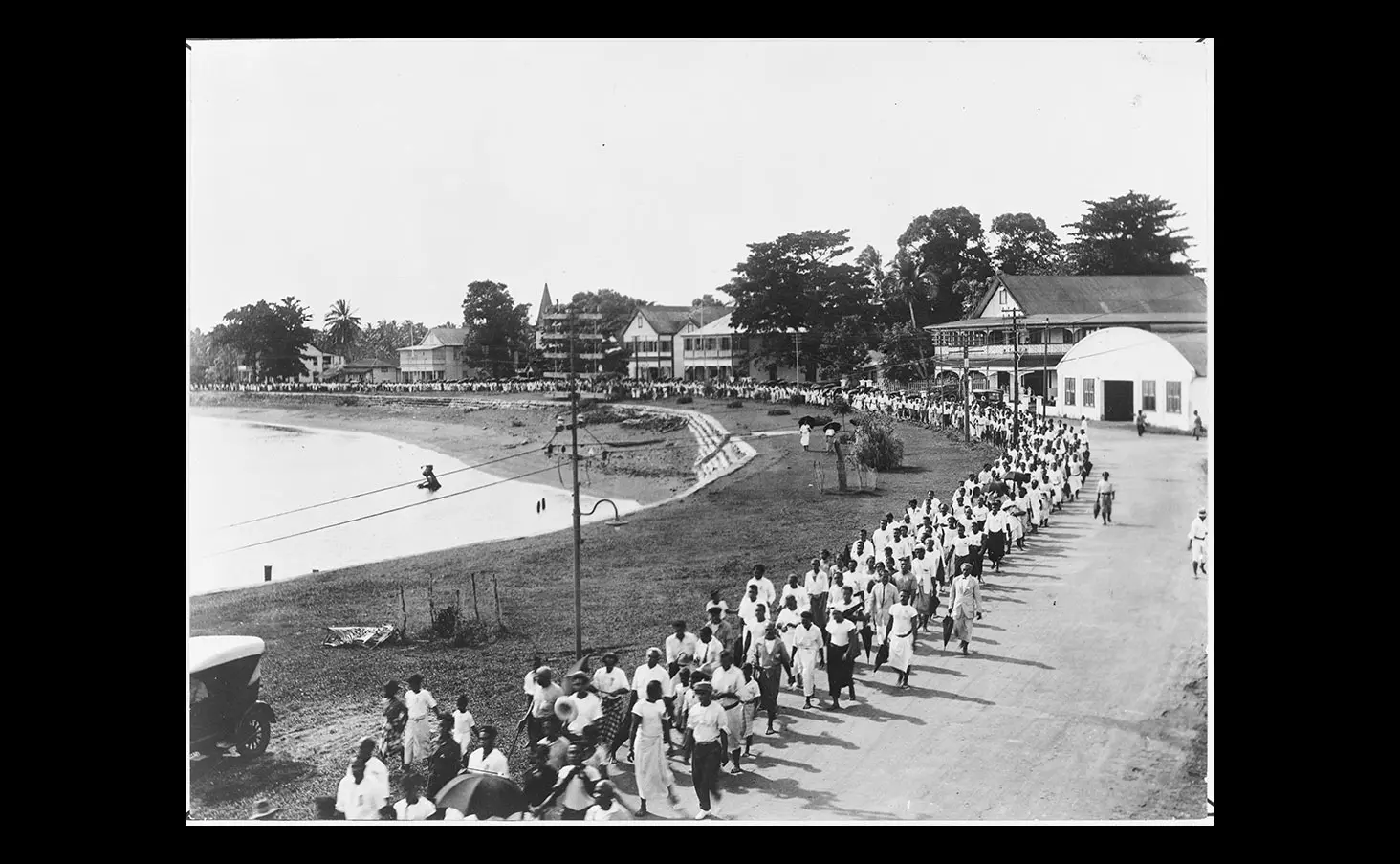 Black and white photo of a Mau movement in Apia — showing Samoan people marching down a road by the coast in Apia, Samoa.