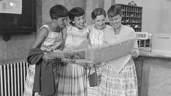 Black and white photo of four smiling young women huddled together as they read a copy of 'The Evening Post' newspaper.