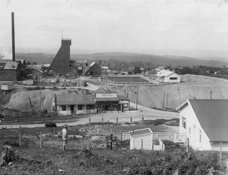 Black and white photo of small township including wooden buildings and large mine headframe and related structures visible in middle distance.