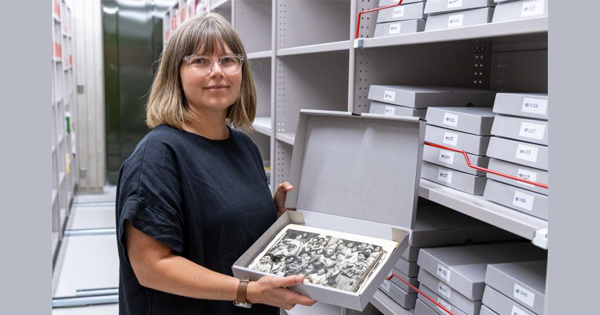 A woman stands among archival shelving lined with boxes of photographic prints while holding open a box and displaying a black and white print.  