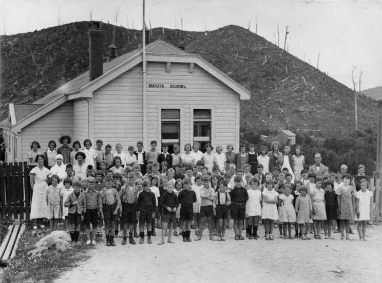 Black and white photo showing large group of students and teachers out front of a small wooden school building with bare hills in the background.