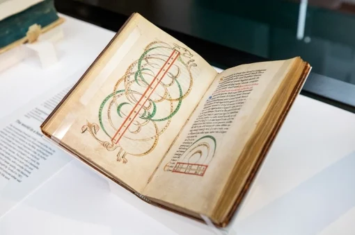 An old book is open to a page with colourful, circular designs, viewed through a glass case. 