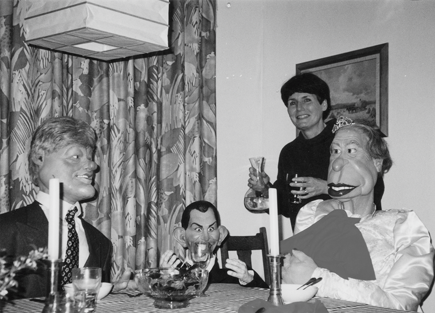 Black and white photo of Susan Foster standing with a wine glass behind a table where three puppets are seated, including Bill Clinton and Queen Elizabeth II.