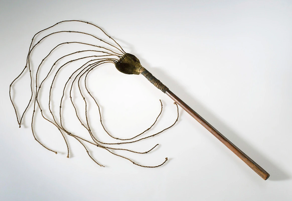 Colour photograph of a cat-o-nine tails whip showing 9 knotted cords attached to a wooden handle.