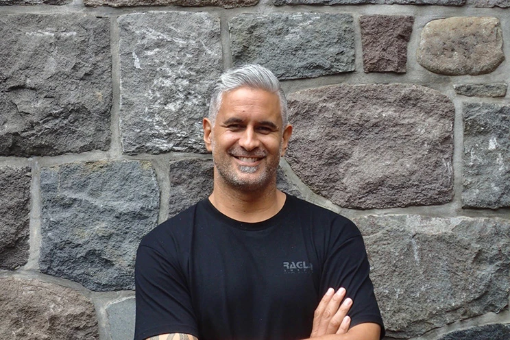 Jay Ruka wearing a black t-shirt with a Raglan surf logo, standing against a stone wall.