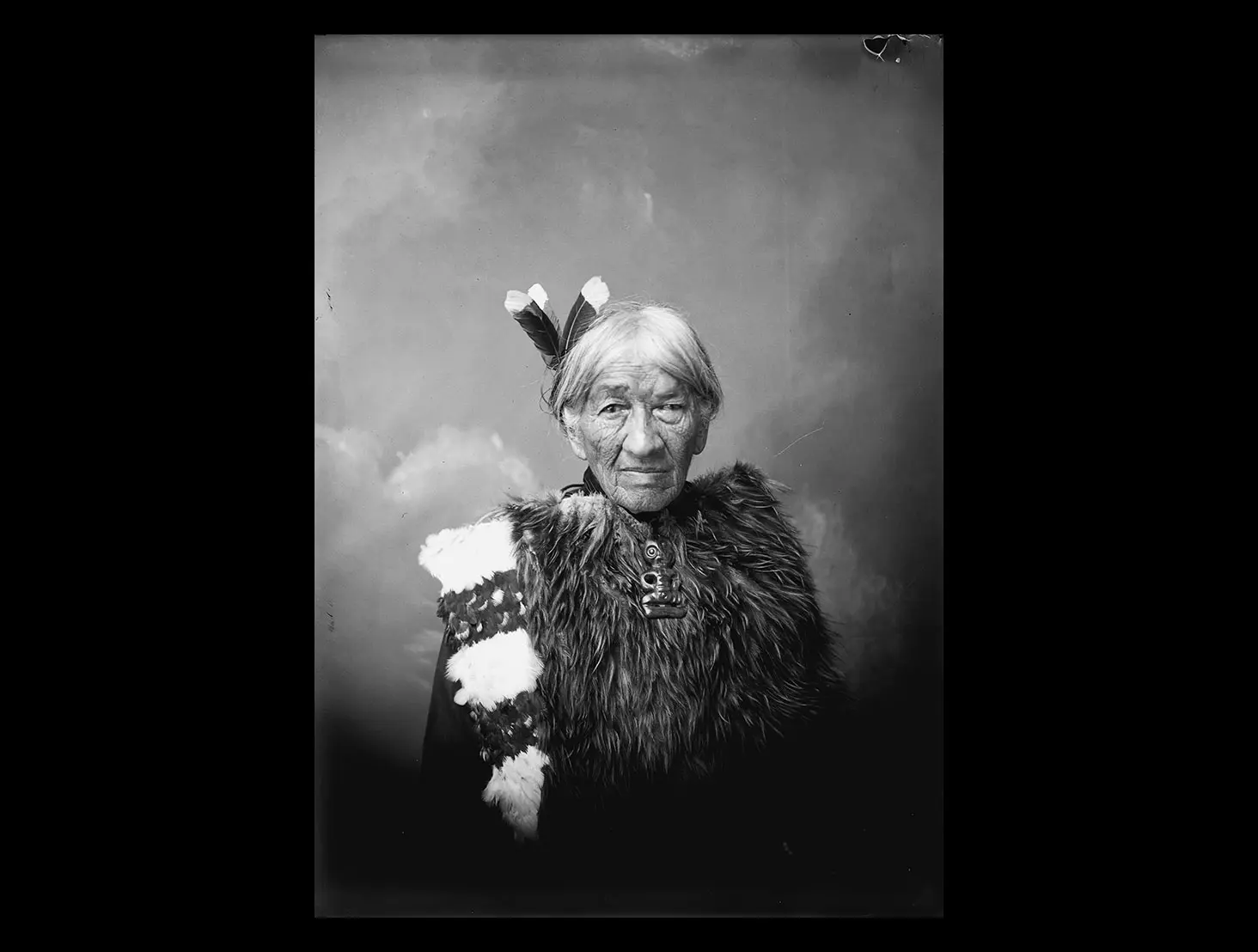 Black and white studio photo of Hēni Te Kiri Karamū wearing a kahu huruhuru (feather cloak), a hei tiki (neck ornament) and 3 huia feathers in her hair.