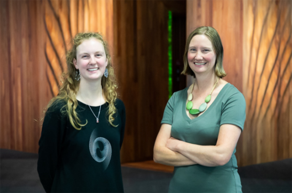 Two smiling women standing in front of a spectacular curved wooden wall. 