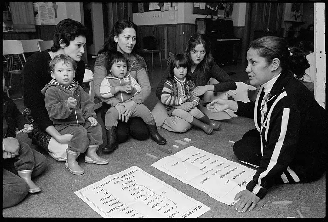 Billie Tait-Jones teaching te reo Māori to a group of mothers and their children. Beside Billie are 2 sheets with kupu Māori.