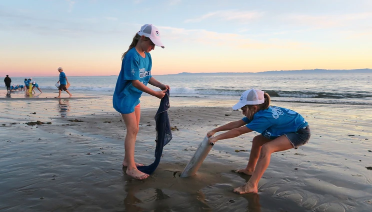 Two primary school students on a beach participating in a science project using ocean research equipment on the sand at sunset with water and other figures in the background