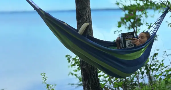 Young girl lying in a hammock reading a book by a lake or the sea.