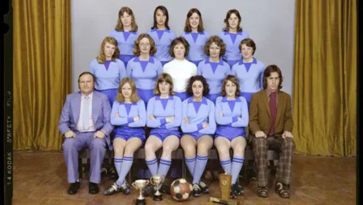 A colour studio portrait of a women's soccer team wearing blue uniforms in three rows, with the captain in middle row wearing white jersey and two male coaches seated at either end of the bottom row. 