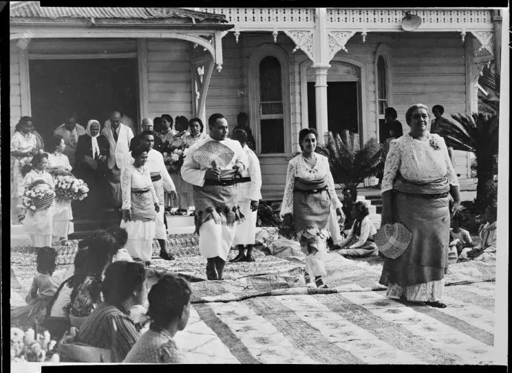 A black and white photograph showing a wedding ceremony with men and women in traditional dress seated and standing on tapa and woven mats. 