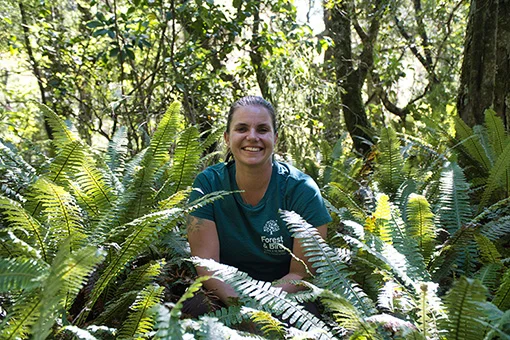 A smiling woman wearing a green t-shirt with the Forest & Bird logo crouching among ferns in the bush.