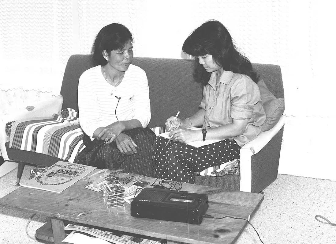 Black and white photo of 2 women from Cambodia — Niborom Young interviewing Sokhom You for the Cambodian oral history project. You has a small microphone connected to a tape recorder on a table close by.