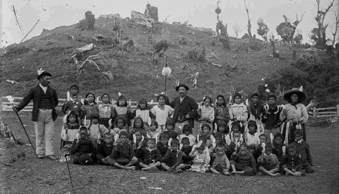 Children from Parihaka posing for a photo, some wearing white feathers in their hair, with Taare (Charles) Waitara standing centre back.