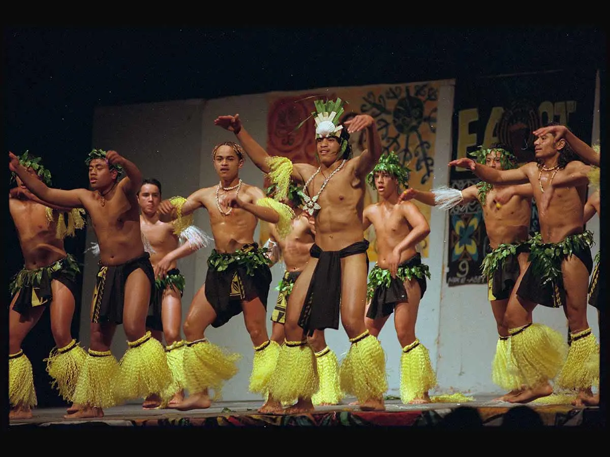 College students in a Rarotongan group performing on stage. Their costumes include decorative headwear, necklaces, black cloth around their waists, and yellow frilled decorations around their lower legs and upper arms.